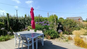 Um pátio externo com mesa de vidro e cadeiras brancas, cercado por vegetação exuberante e flores roxas.