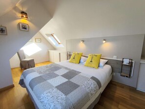 A bedroom with a sloped ceiling, a bed with patterned bedding, two pillows, a chair, and wall-mounted lighting.