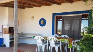 Een terras buiten met een tegelvloer, witte plastic stoelen, een tafel met een groen-geel geruit tafelkleed, een pizzaoven en potplanten.