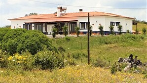 Een gelijkvloers huis met een rode dakbedekking, omringd door weelderig groen en een veld vol gele bloemen.