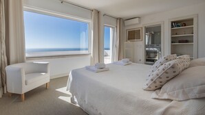 A bedroom with a large window, a bed with white bedding, a white armchair, and a shelf with books. - Luxury Beachfront Villa (Colares)