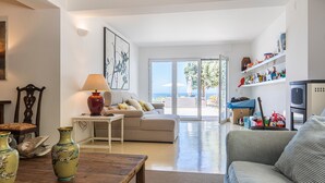 A spacious living room with a sofa, a coffee table, and decorative vases. There is a sliding glass door leading to a balcony with a view of the sea. - Luxury Beachfront Villa (Colares)