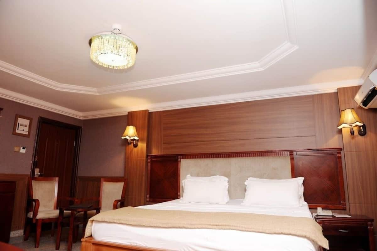 A hotel room with a large bed, wooden headboard, two bedside lamps, a chandelier, and a wooden wardrobe.