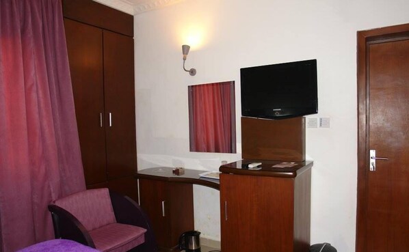 A compact room with a wooden wardrobe, a small desk with a flat-screen TV, a pink chair, and a red curtain.