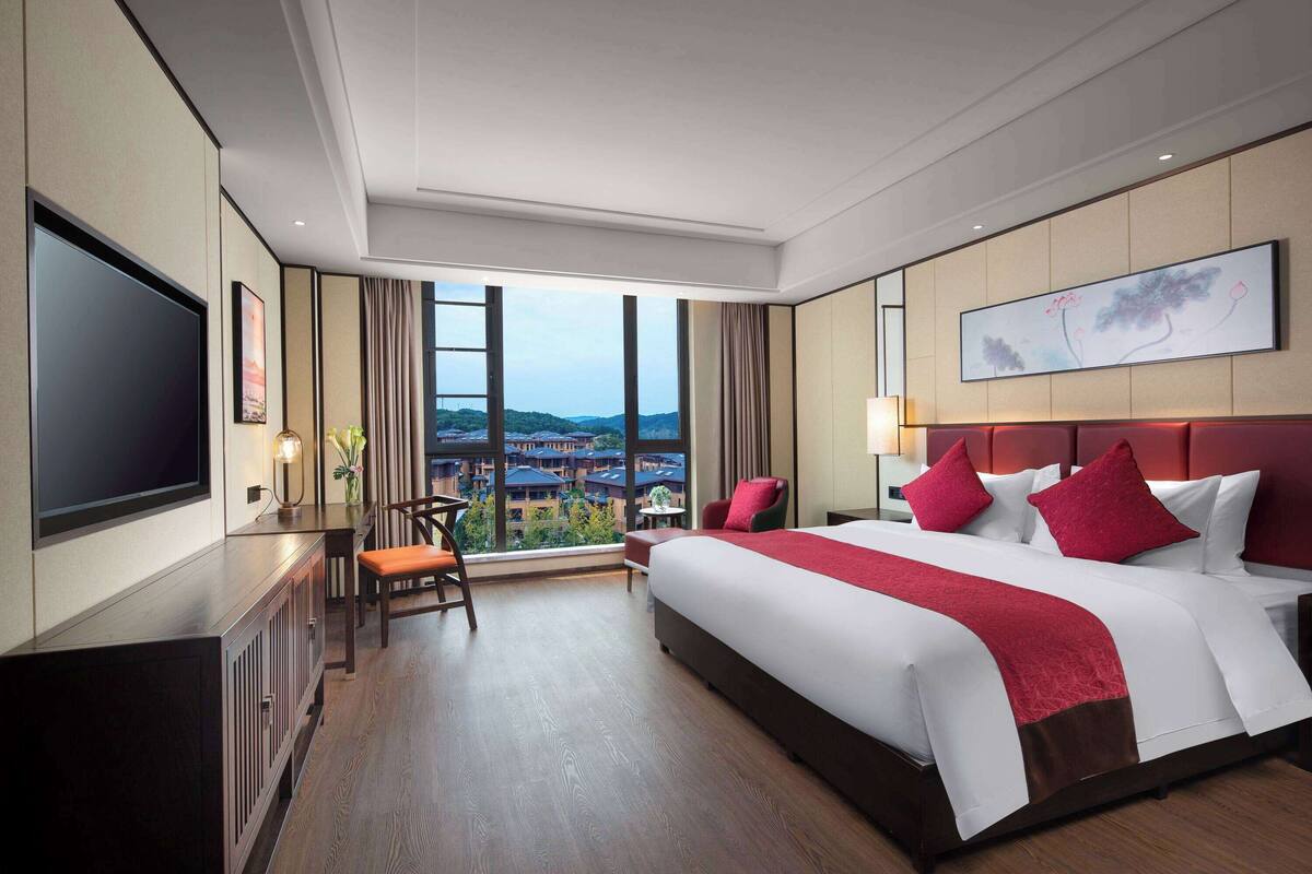 Ein Hotelzimmer mit einem großen Bett, einem Flachbildfernseher, einem Schreibtisch und Blick auf eine Stadt durch große Fenster.