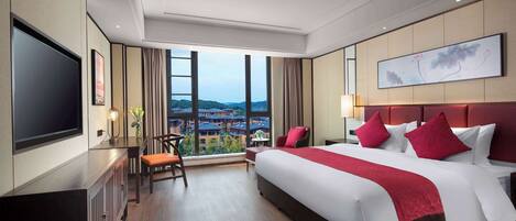 A hotel room with a large bed, a flat-screen TV, a desk, and a view of a cityscape through large windows.