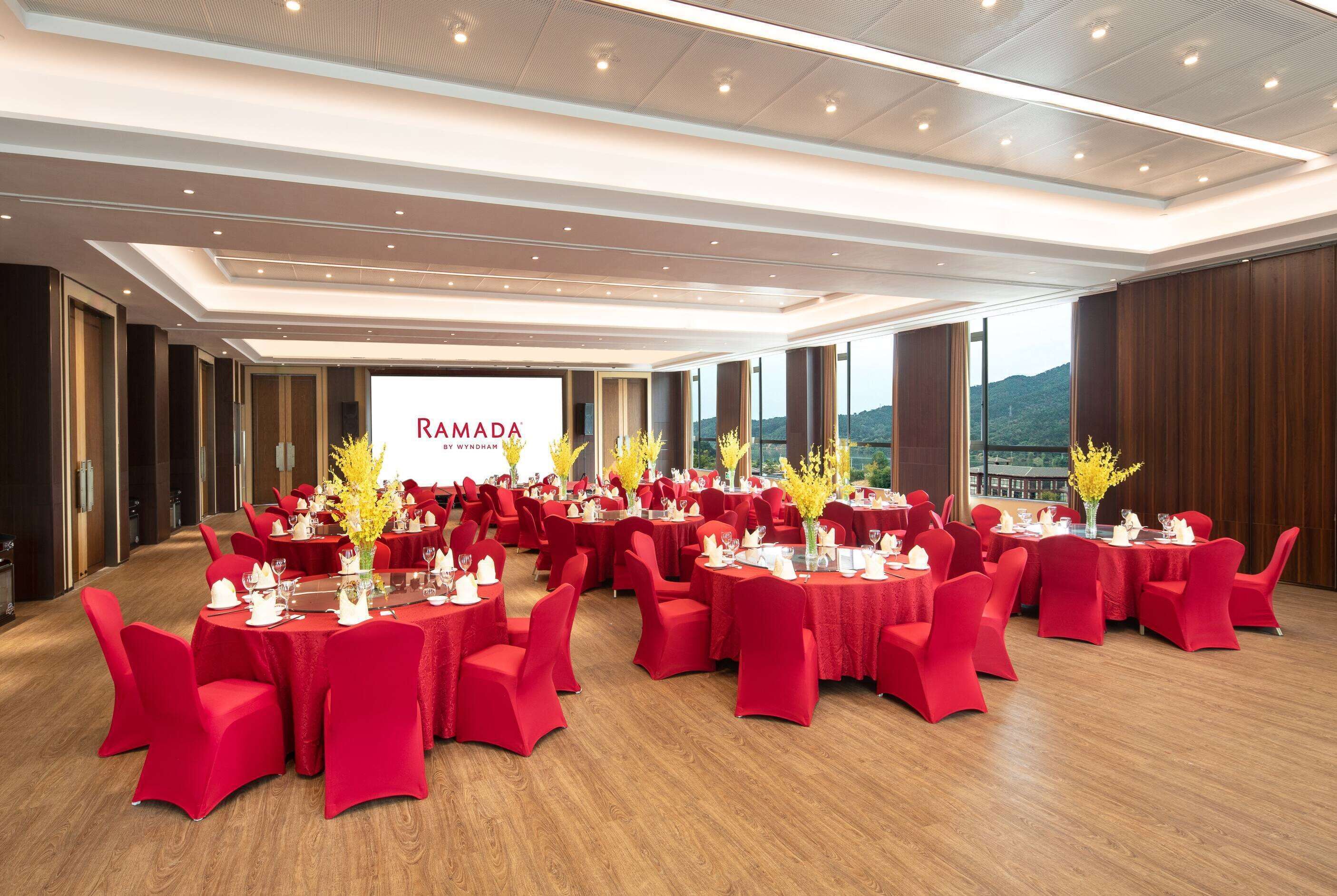 Ett konferensrum med flera bord och stolar, en stor skärm som visar texten ”RAMADA”, och en utsikt över ett bergigt landskap genom stora fönster.