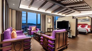 Una habitación de hotel moderna con un ventanal grande, un conjunto de sofás, un televisor de pantalla plana y una cama con almohadones rojos.