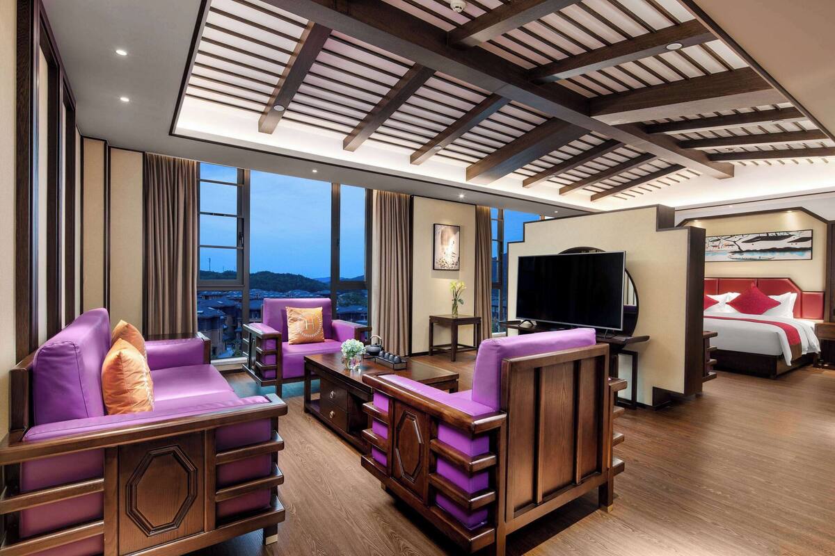 Ein modernes Hotelzimmer mit großem Fenster, einer Sitzecke, einem Flachbildfernseher und einem Bett mit roten Kissen.