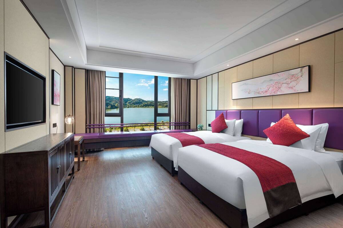 Ein Hotelzimmer mit zwei Betten, einem Fernseher, einem Schreibtisch und einem großen Fenster mit Seeblick.