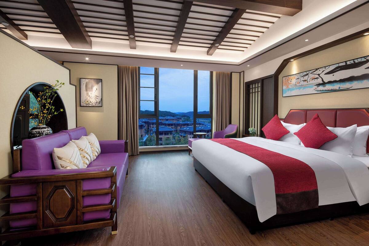 Ein Hotelzimmer mit einem großen Bett, einem violetten Sofa, Blick auf die Stadt durch große Fenster und einem modernen Deckenstil.