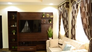 A living room with a large flat-screen TV mounted on a wooden entertainment unit, a beige sofa with decorative pillows, and a potted plant. - Marshall Hotel Apartments (Kano)
