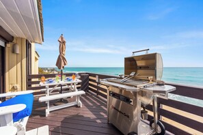 An outdoor dining area with a grill, tables, and chairs, overlooking the ocean.