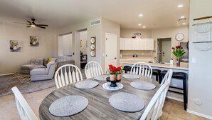 A modern kitchen with white cabinets, a dining area with a round table, and a living room with a sofa and a ceiling fan. - Group Luxury Retreat ~ 4 Neighboring Homes, 21 Beds, Pool & Golf Community (Mesa)