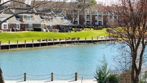 A riverside view of a residential area with apartment buildings, a wooden dock, and a grassy lawn.