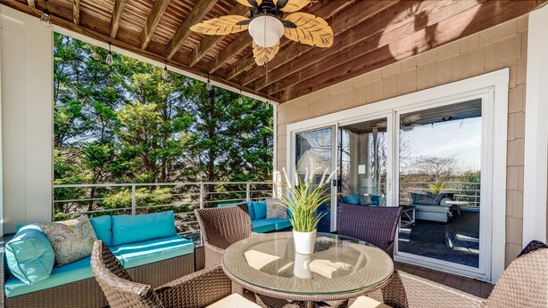 A covered outdoor seating area with wicker furniture, a glass table, and a ceiling fan.