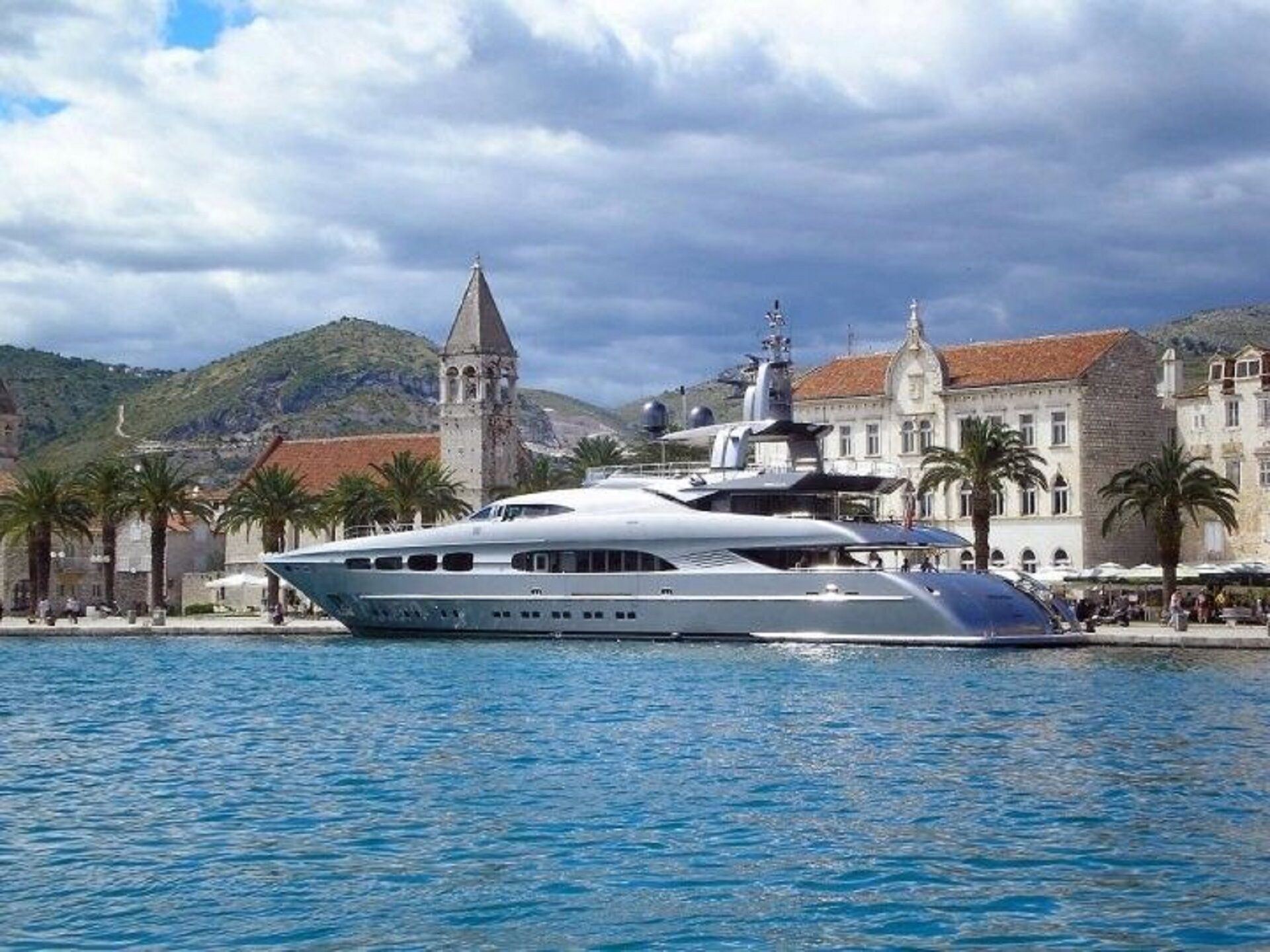A large yacht docked near a historic town with a prominent church tower and traditional architecture.