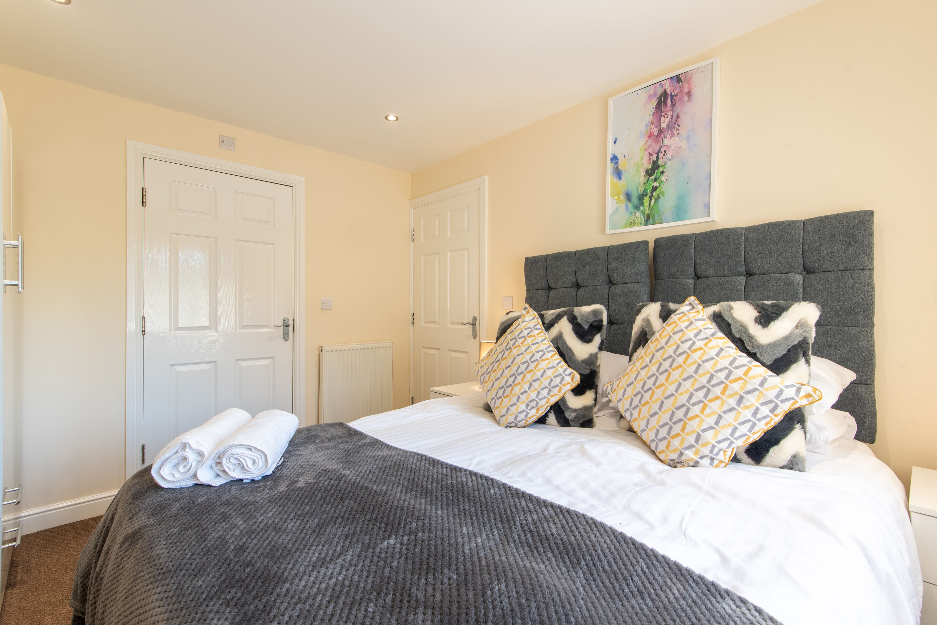 A neatly made bed with a grey headboard, two patterned pillows, and rolled towels on the side.