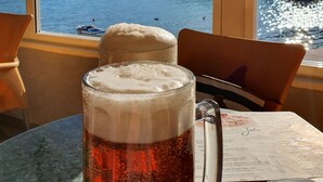 Ein Bierglas mit Schaumkrone auf einem Tisch, neben dem eine Speisekarte liegt, mit Blick auf ein Gewässer und Gebäude im Hintergrund.