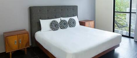 Un letto grande con una testiera grigia imbottita, biancheria da letto bianca e cuscini decorati. Sono presenti due comodini in legno con cassetti e un'ampia finestra che si affaccia sul verde.