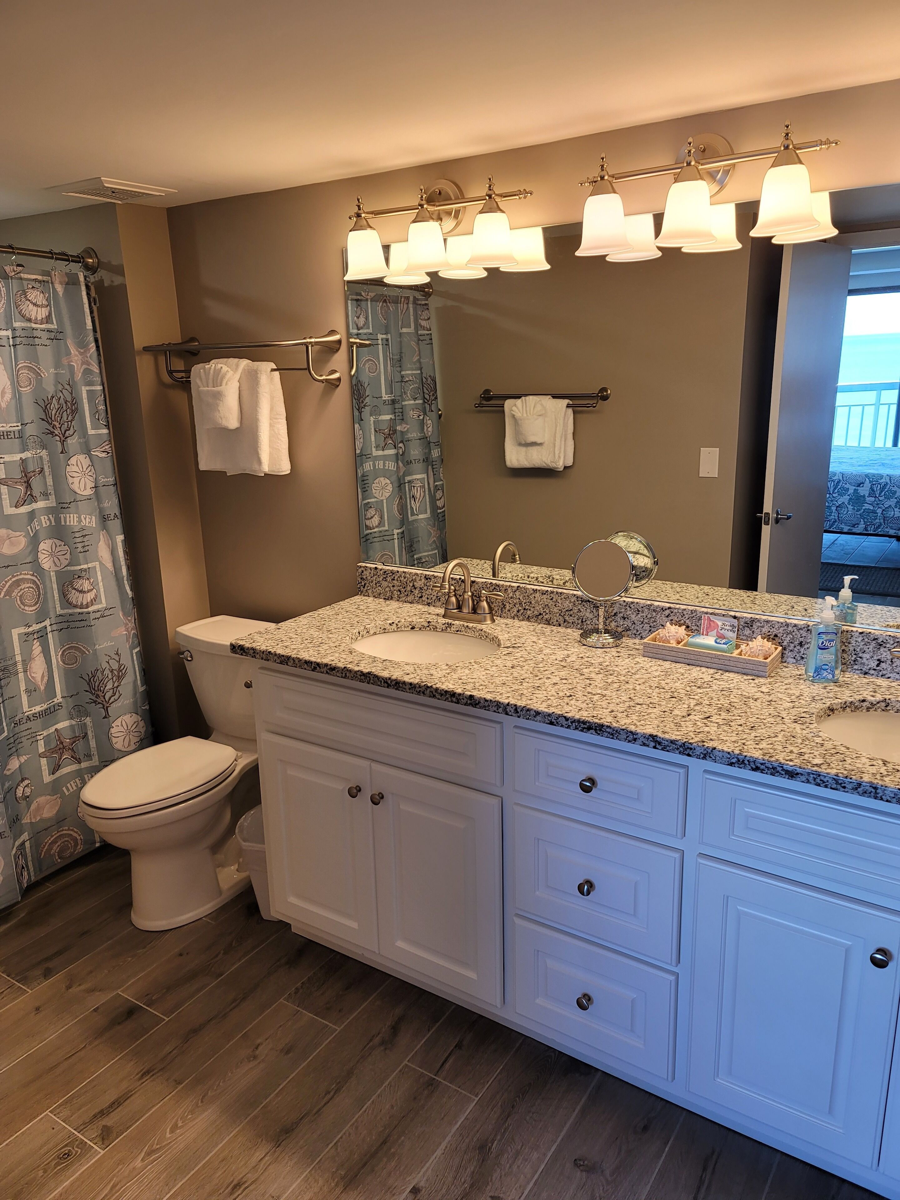 A bathroom with a granite countertop, a white toilet, a vanity with drawers, and a shower curtain with a sea shell pattern.