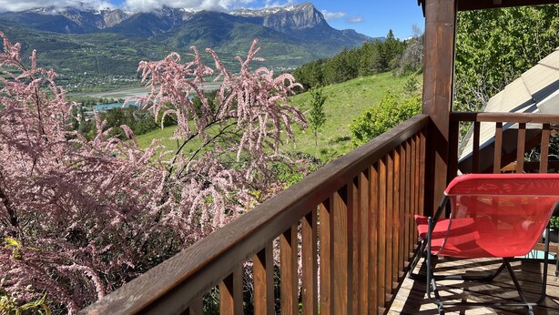 Terraza de madera con una silla roja, con vistas a un paisaje montañoso con árboles en flor y cielo despejado.
