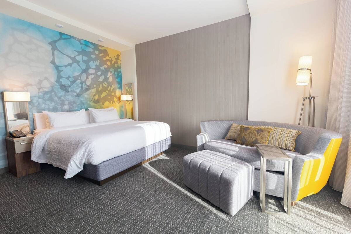 Une chambre d’hôtel moderne avec un grand lit, un fauteuil gris, une petite table et une fresque murale abstraite et colorée.