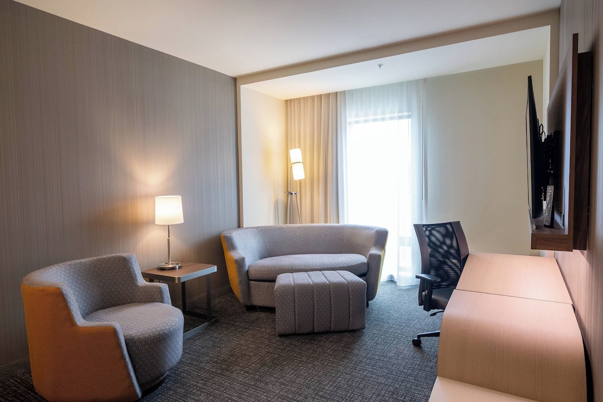 Une chambre d’hôtel équipée d’un canapé, d’un fauteuil, d’un pouf, d’un bureau, d’une lampe et d’un téléviseur posé sur un meuble.