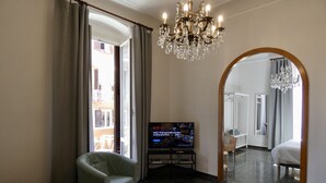 Habitación de hotel con televisor, un sillón verde, una mesita de centro, una araña y un espejo grande.