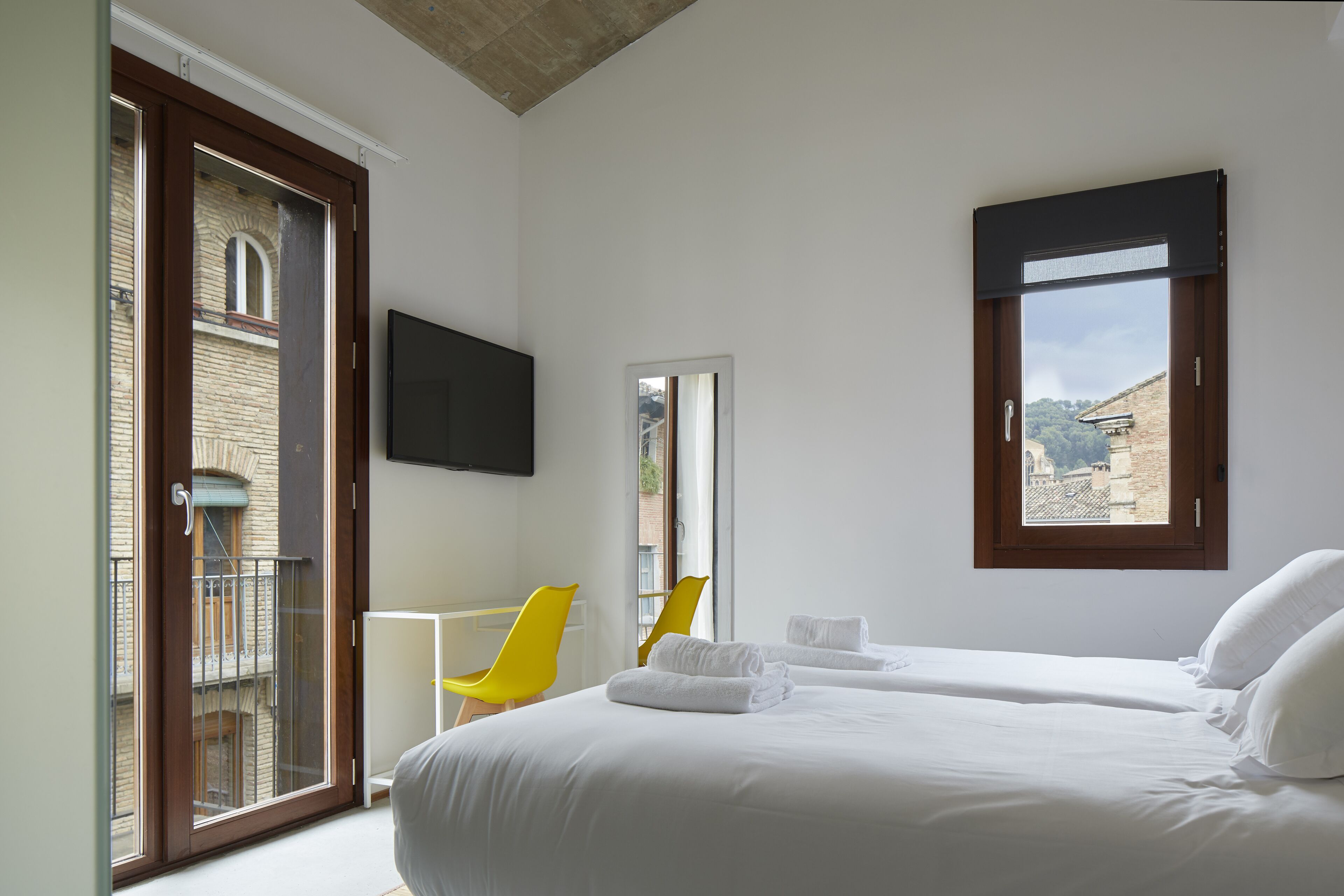 A hotel room with a bed, a TV, a balcony with a chair, and a view of buildings.