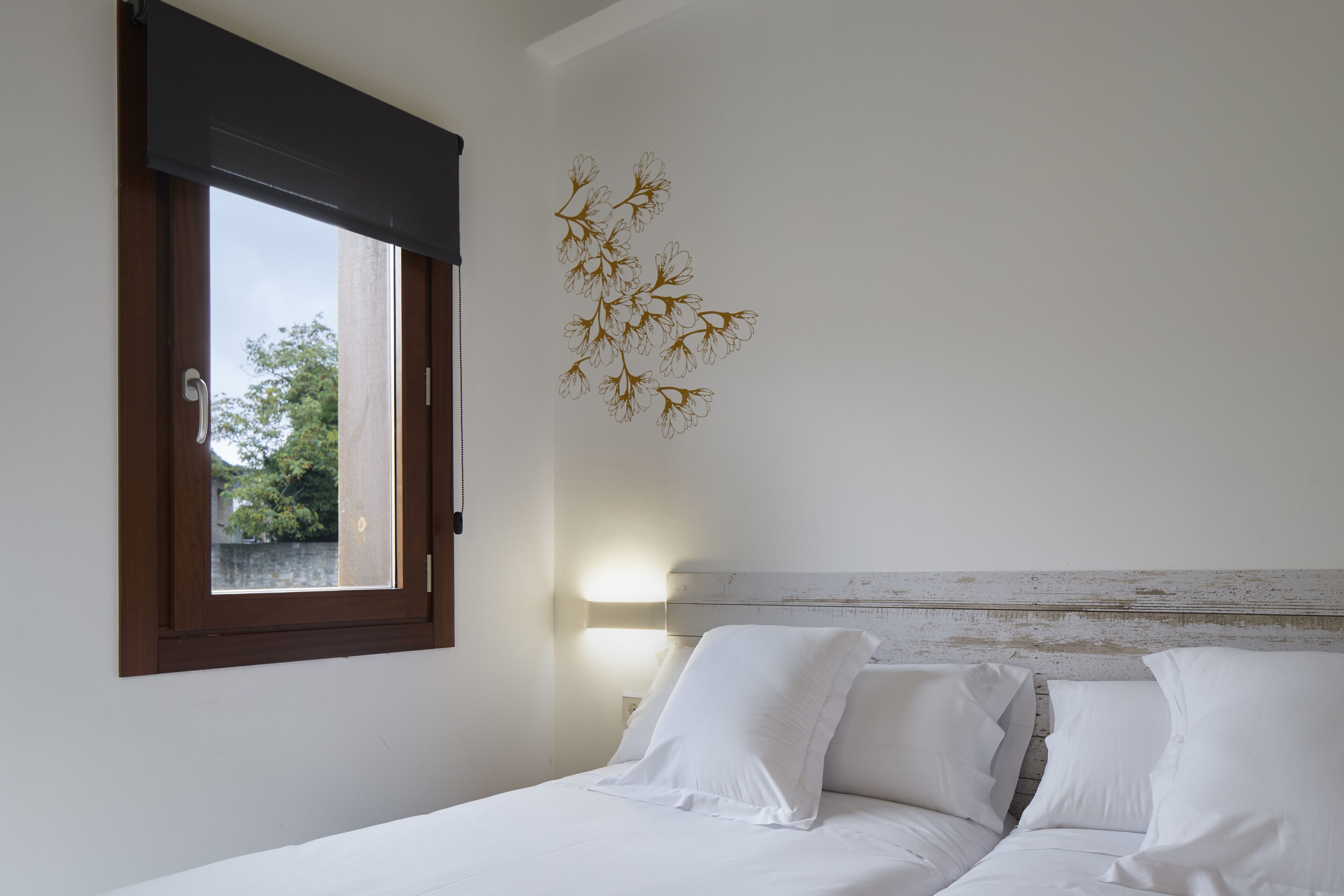 Une chambre à coucher avec un lit, des oreillers blancs, une table de chevet, une lampe fixée au mur et une fenêtre donnant sur des arbres.