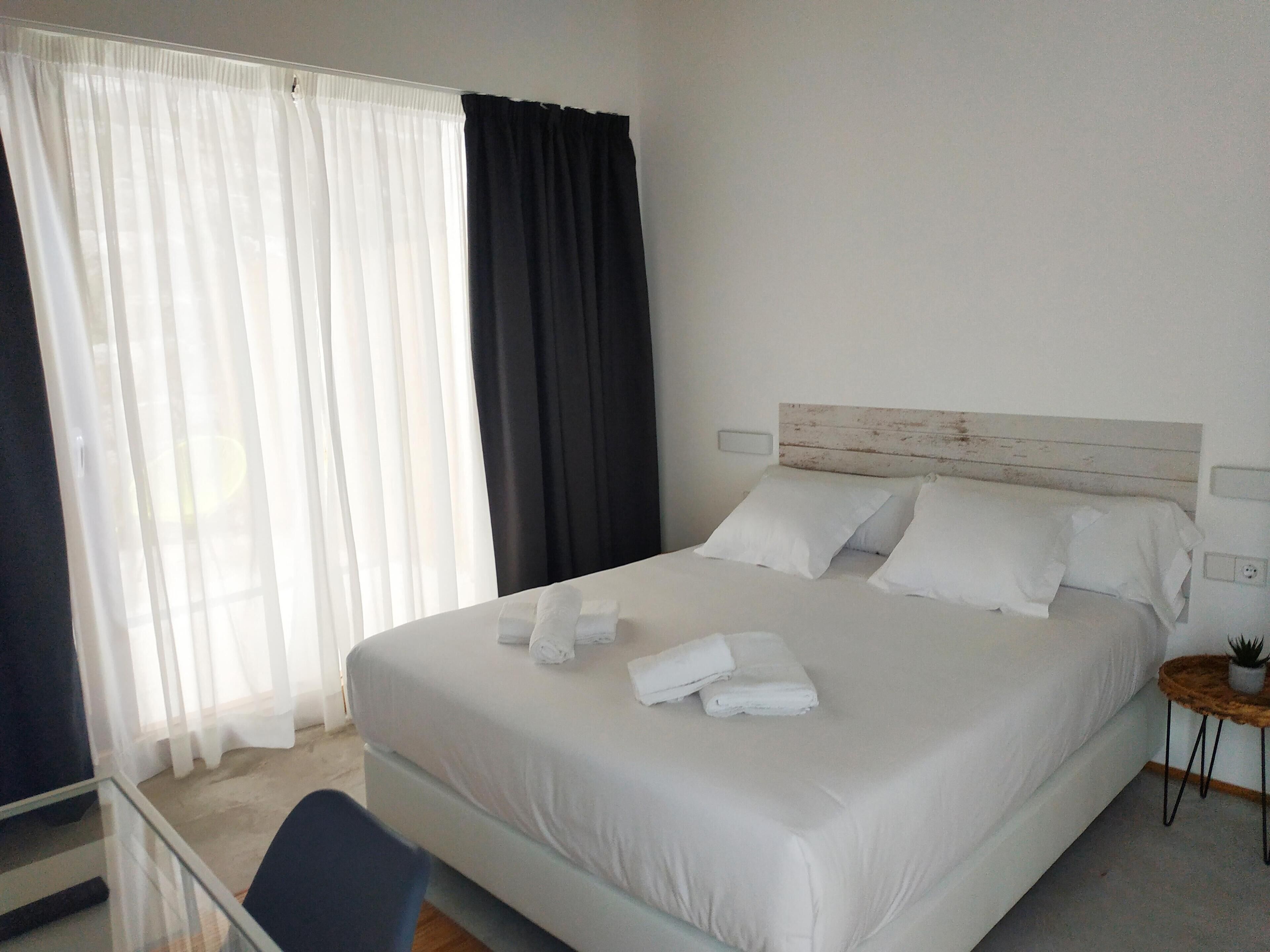 A hotel room with a large bed, white linens, a wooden headboard, a bedside table with a plant, and sheer curtains.