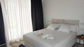 A hotel room with a large bed, white linens, a wooden headboard, a bedside table with a plant, and sheer curtains.