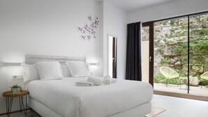 A modern bedroom with a large bed, a small side table, a wall-mounted light, and a view of a stone wall and greenery outside.