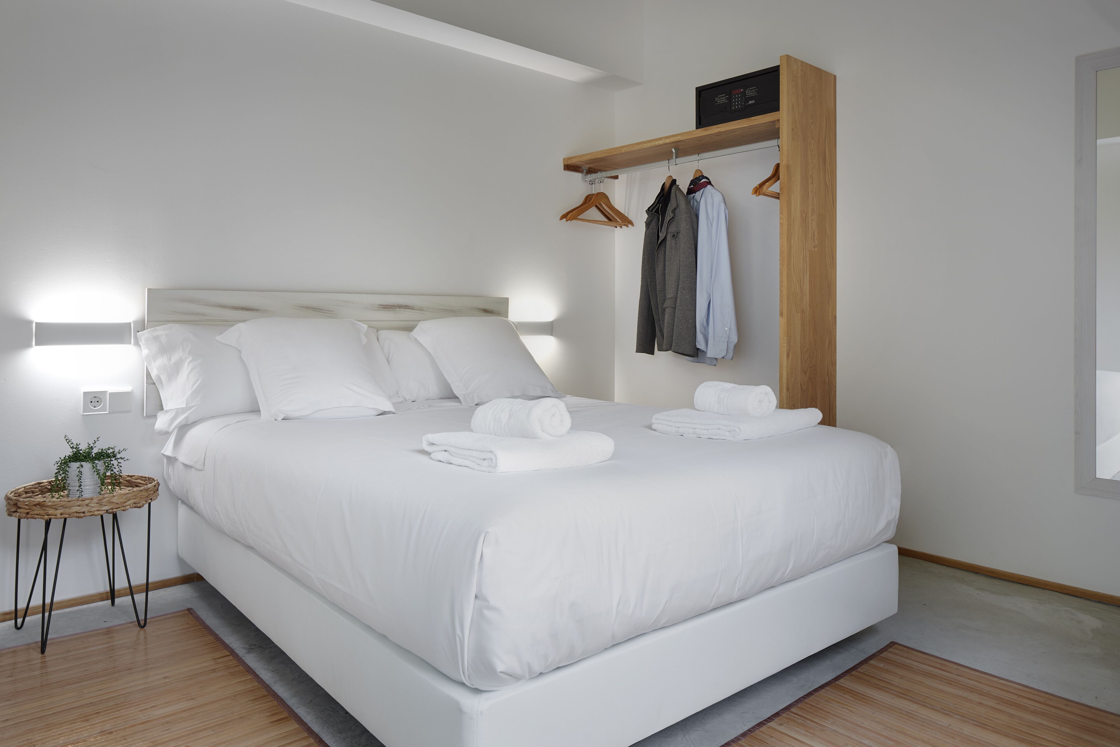 Une chambre moderne avec un grand lit, du linge de lit blanc, une table de chevet agrémentée d’une plante et une étagère en bois où sont posés des vêtements.