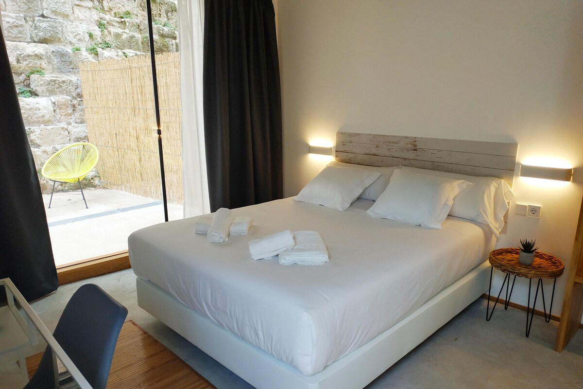 A hotel room with a bed, a bedside table, a chair, and a view of the outside.