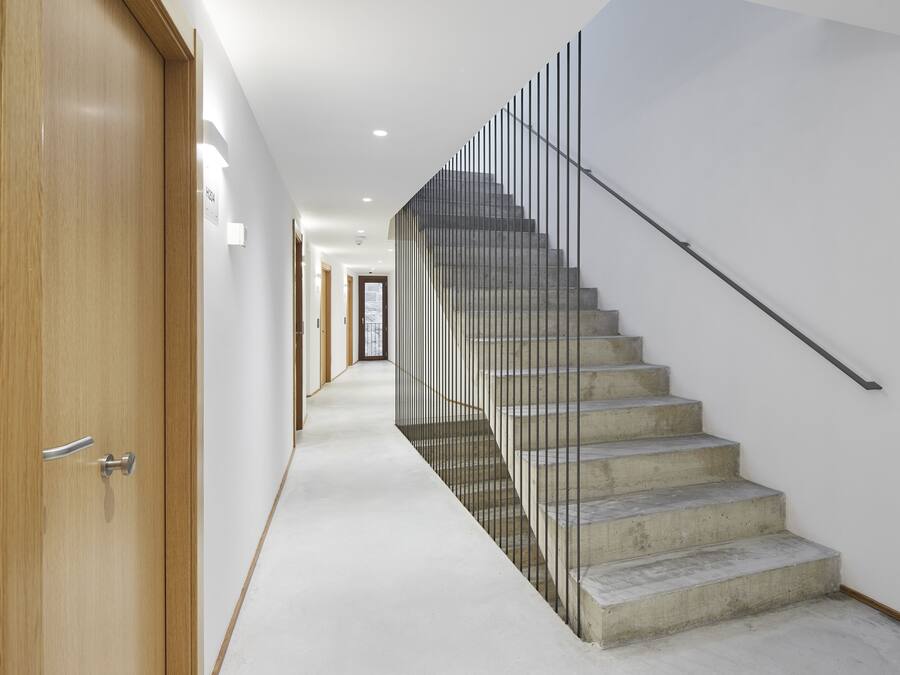 Un interior moderno con una puerta de madera, una escalera de concreto con barandillas metálicas y un pasillo blanco.