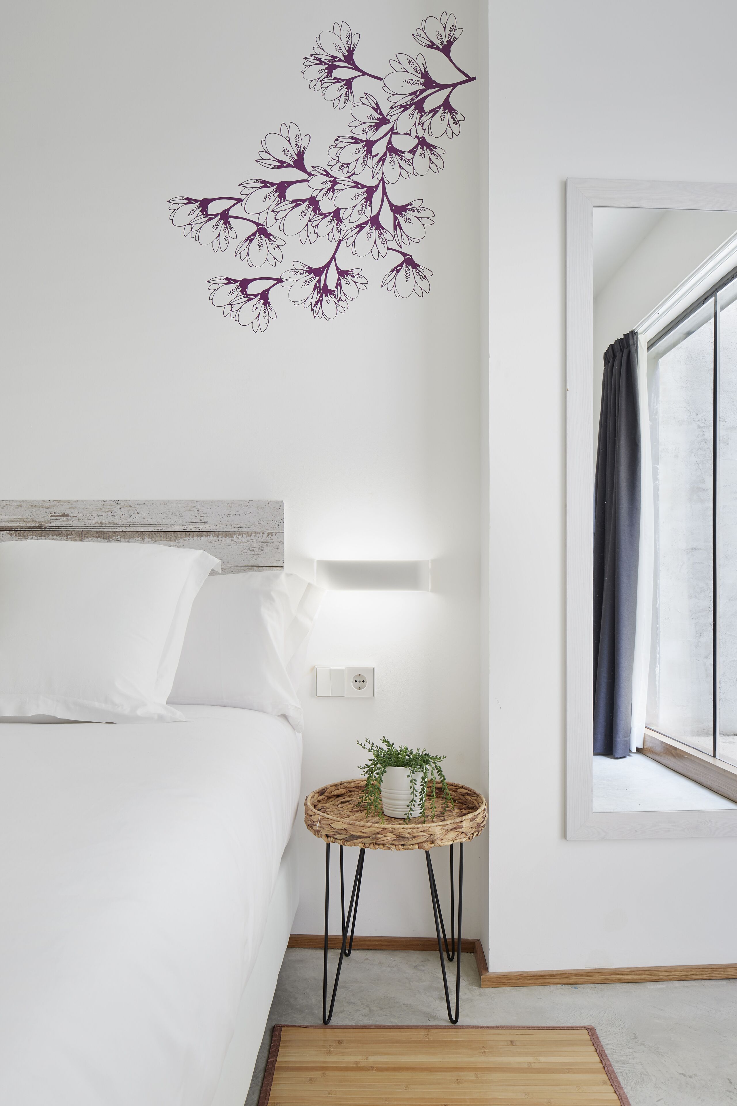A bedroom with a bed, a wall-mounted light, a small side table with a plant, and a window with curtains.