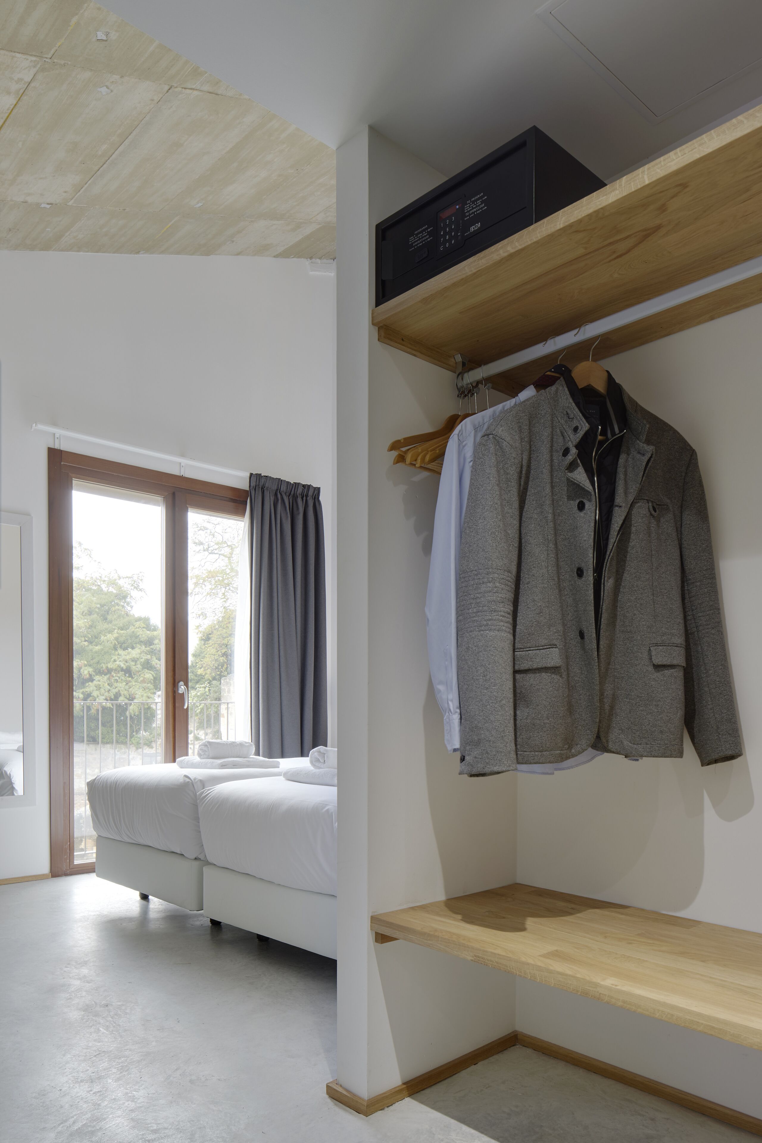 A bedroom with a bed, a wooden ceiling, a wardrobe with clothes, and a window with curtains.