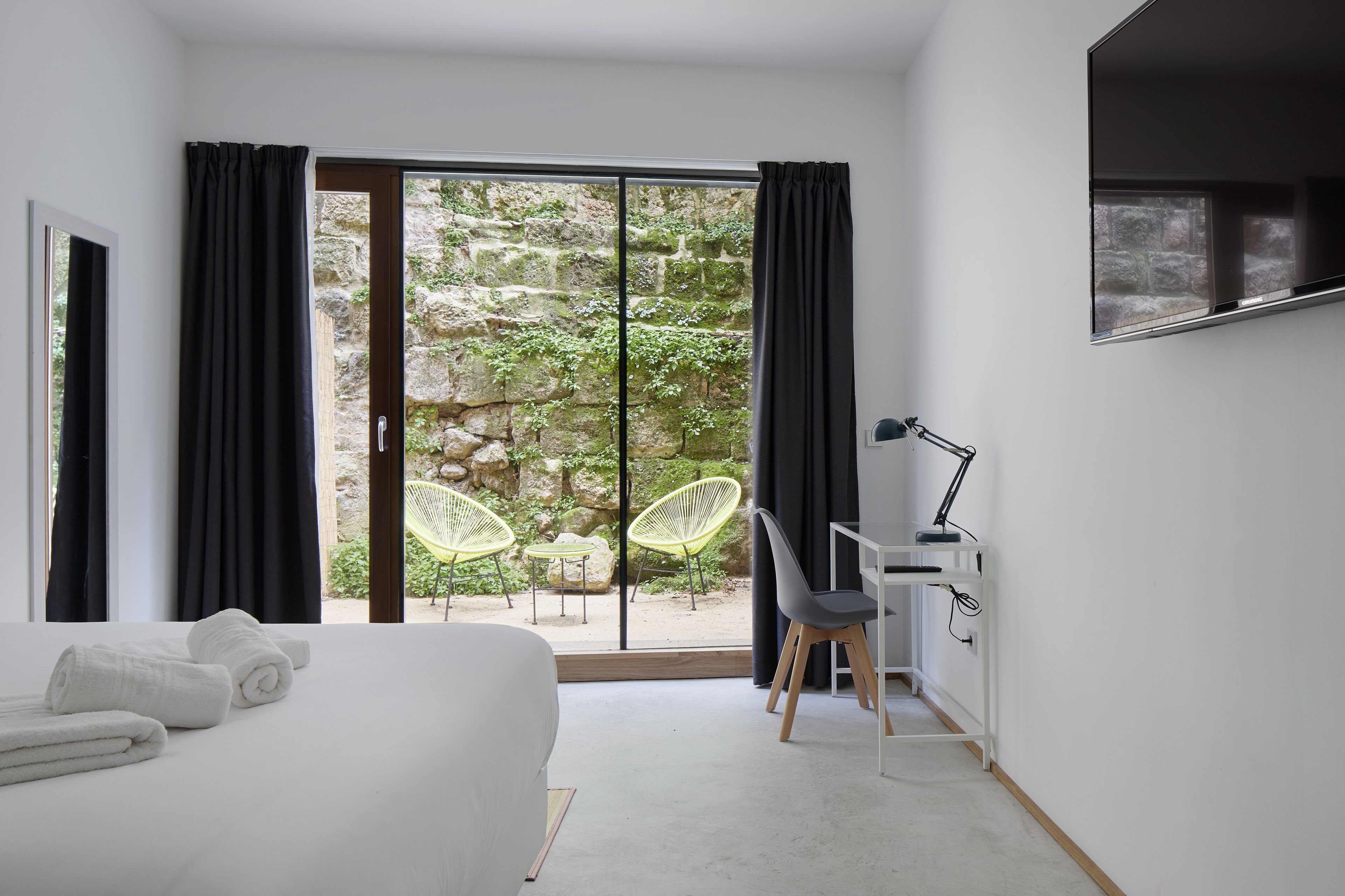 A modern bedroom with a large bed, a desk with a chair, and a view of a garden through a sliding glass door.
