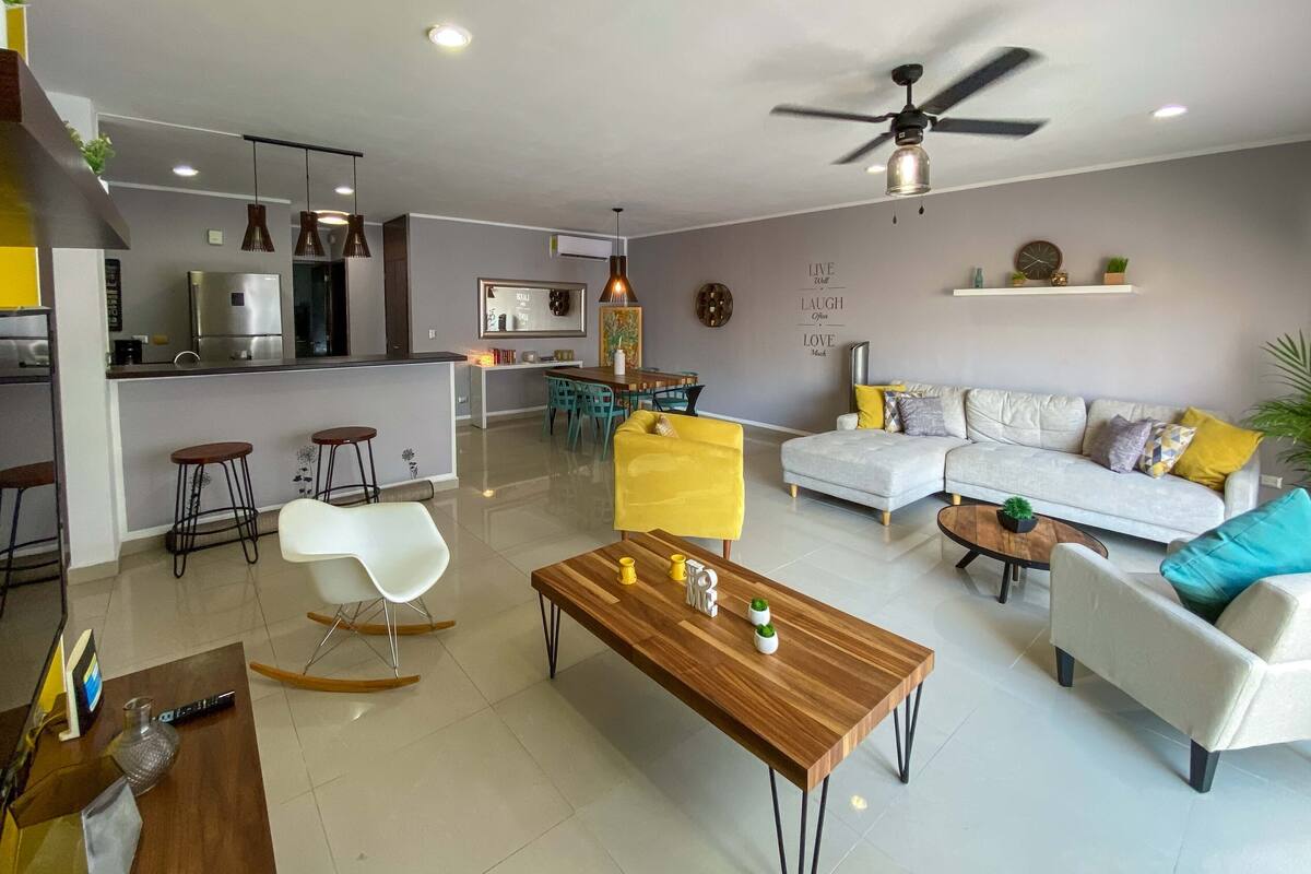 Uma sala de estar moderna com sofá, mesa de centro e cadeiras. Há uma cozinha com geladeira e mesa de jantar.