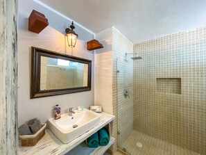 A bathroom with a white sink, a mirror, a shower with beige tiles, and a wooden shelf with towels and toiletries. - Plaza Paraiso (Playa del Carmen)