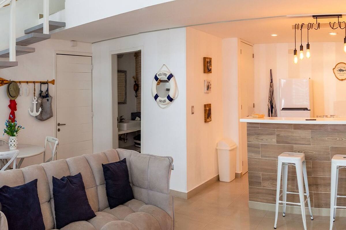 Uma sala moderna com um sofá cinzento, uma mesa de jantar com cadeiras, uma escada e uma área de cozinha com banquetas.