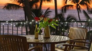 Una mesa de madera con sillas y un jarrón de flores en un balcón con vista a palmeras y al atardecer.