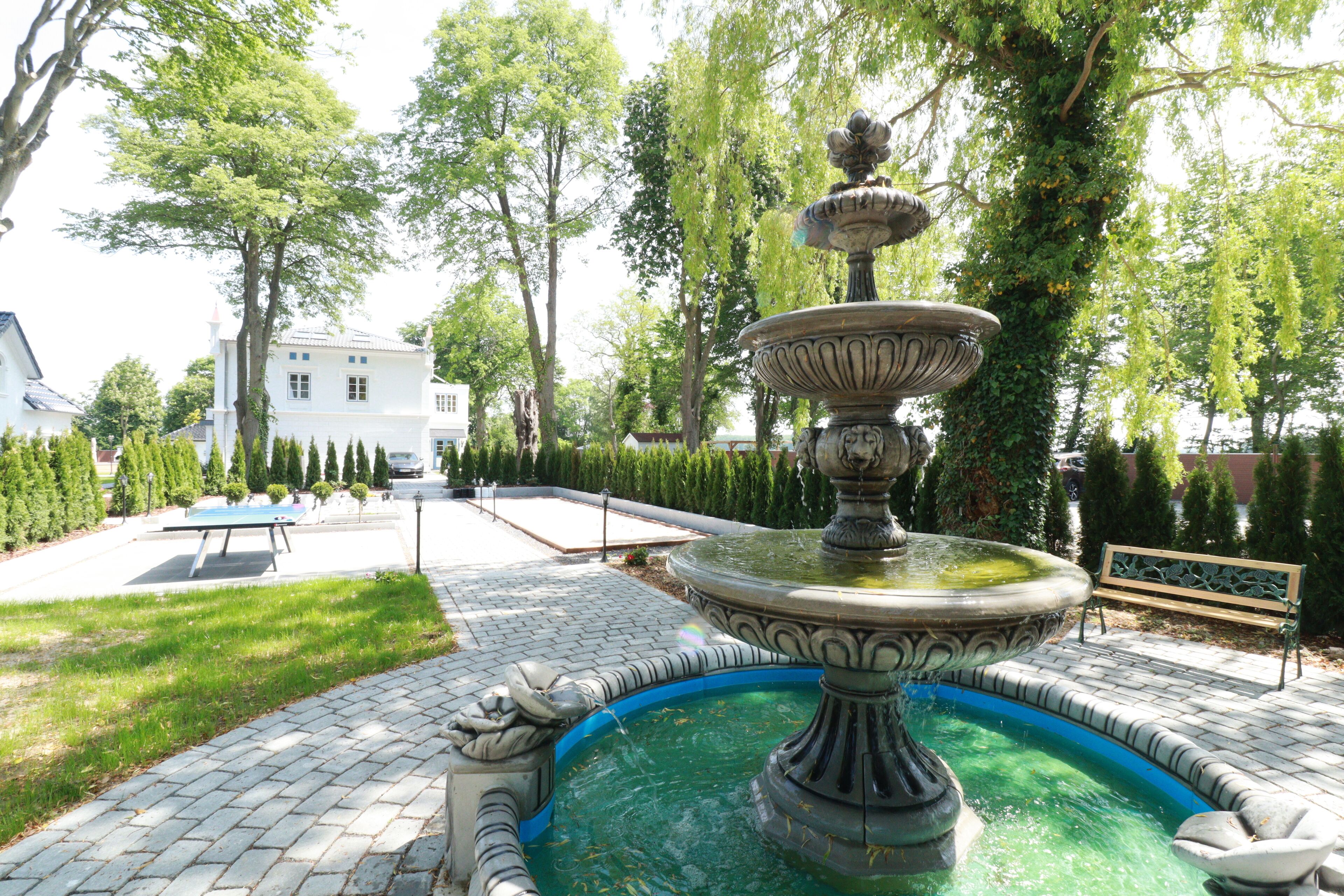 A fountain with a circular basin and a tiered structure, surrounded by a paved area with a bench and a pool in the background.
