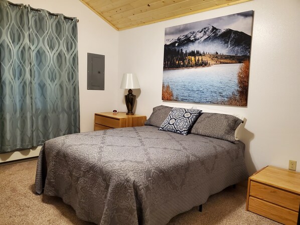 Un dormitorio con cama, mesita de noche, lámpara, un cuadro de un paisaje montañoso y cabecero de madera.