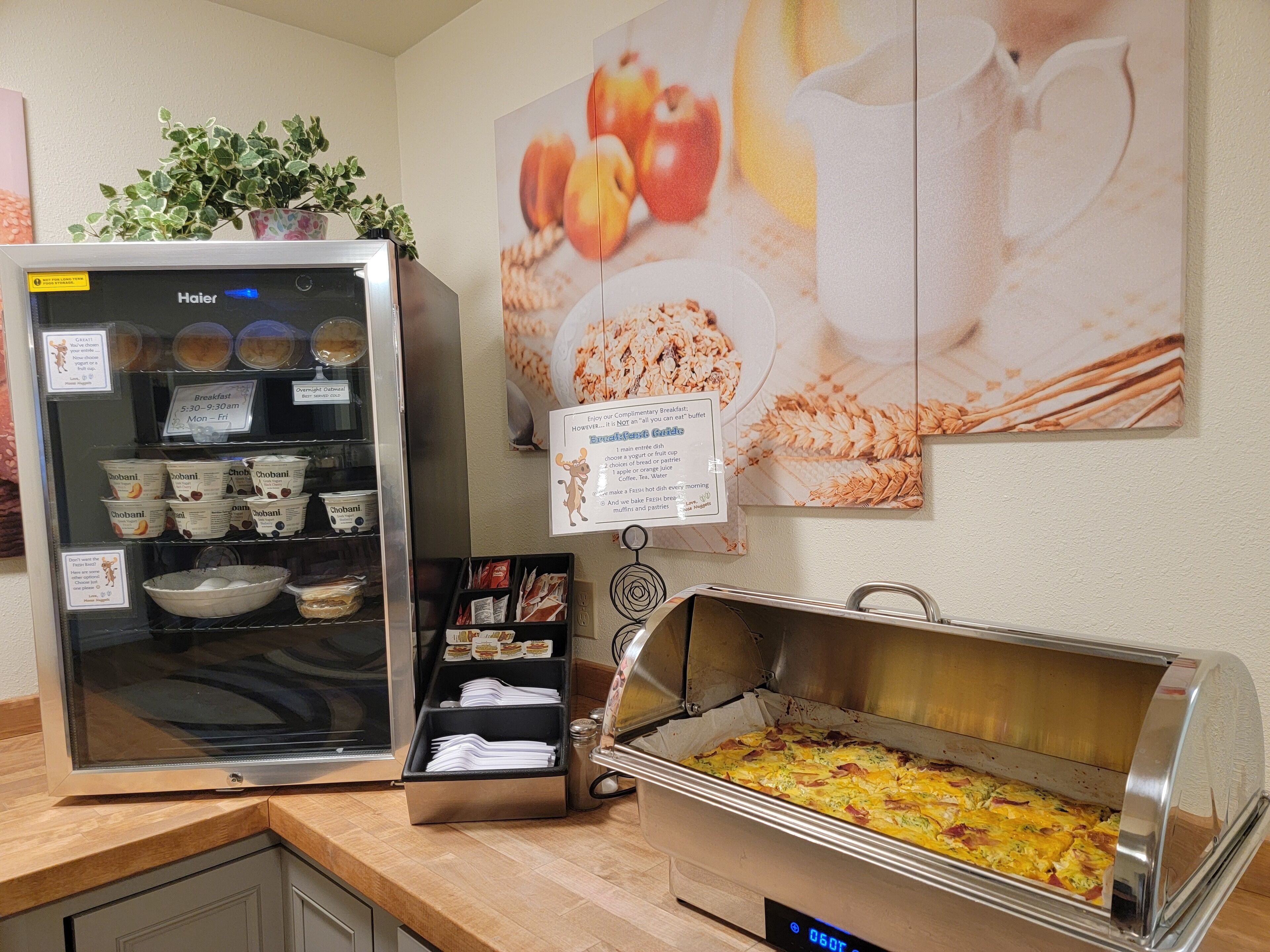 Um buffet de café da manhã com diversas opções de comida, uma geladeira com bebidas e uma parede com imagens de frutas e cereais.