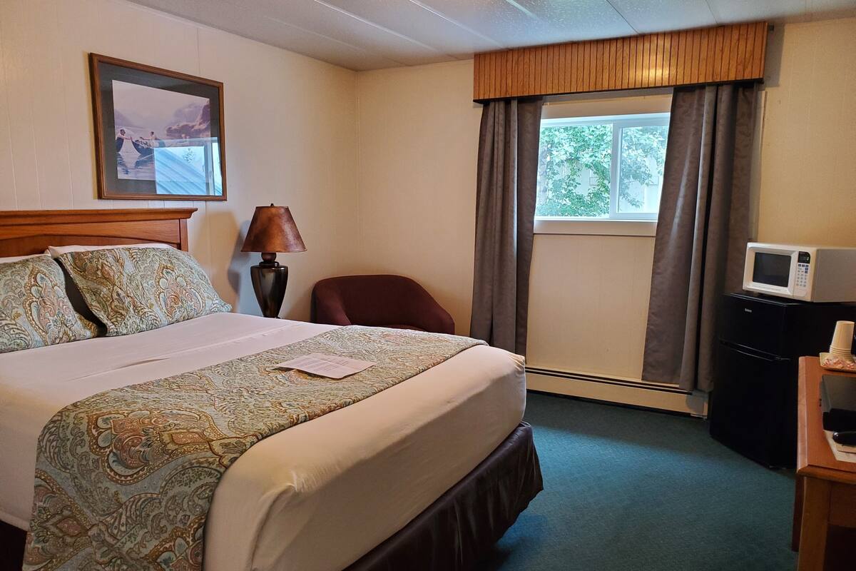 A hotel room with a large bed, a desk with a microwave, a chair, a lamp, and a window with curtains.