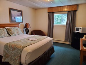 A hotel room with a large bed, a desk with a microwave, a chair, a lamp, and a window with curtains.