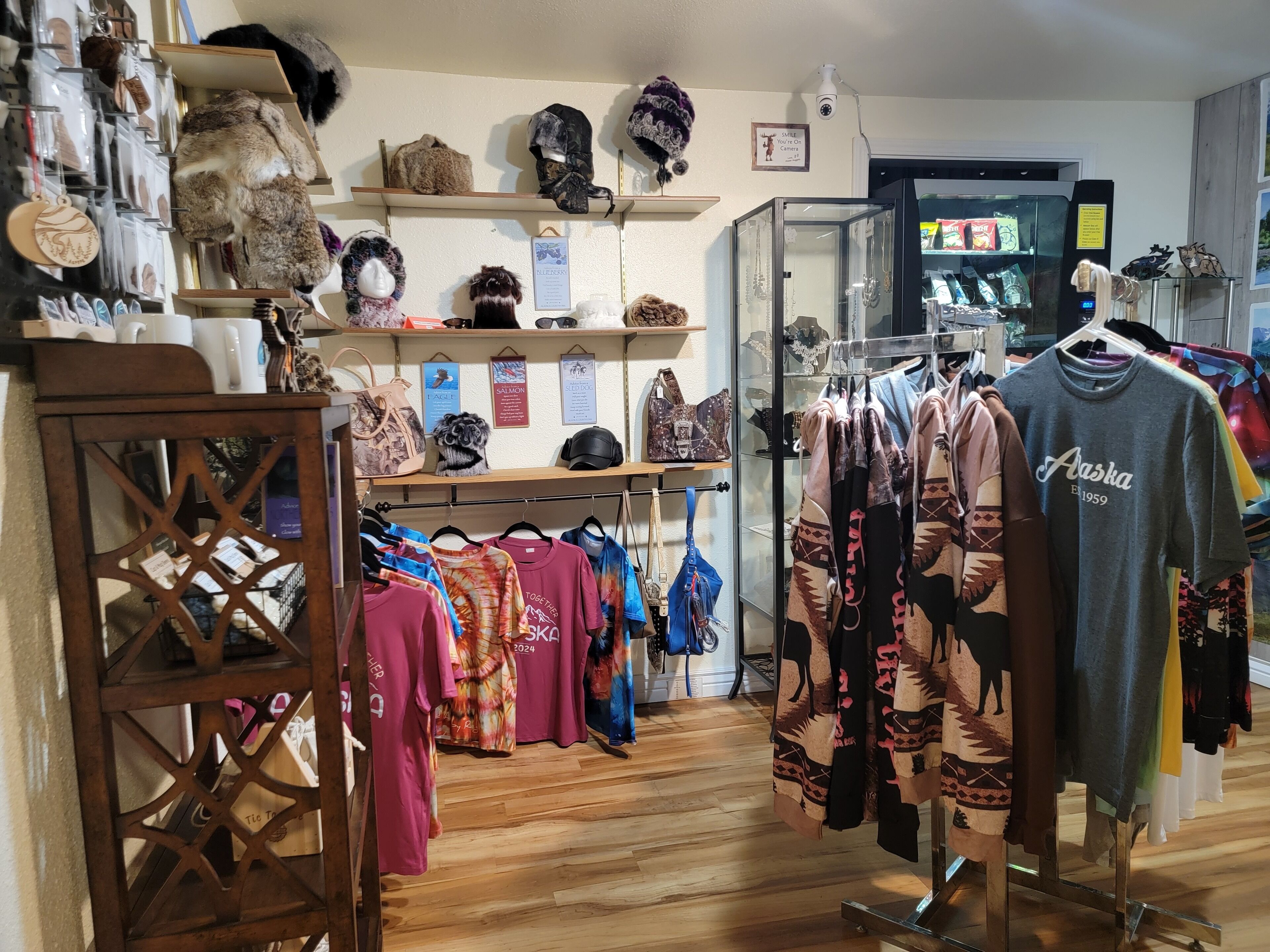 Una tienda minorista con estantes que exhiben sombreros y bolsos, perchas con ropa colorida, y una cámara de seguridad en la pared.